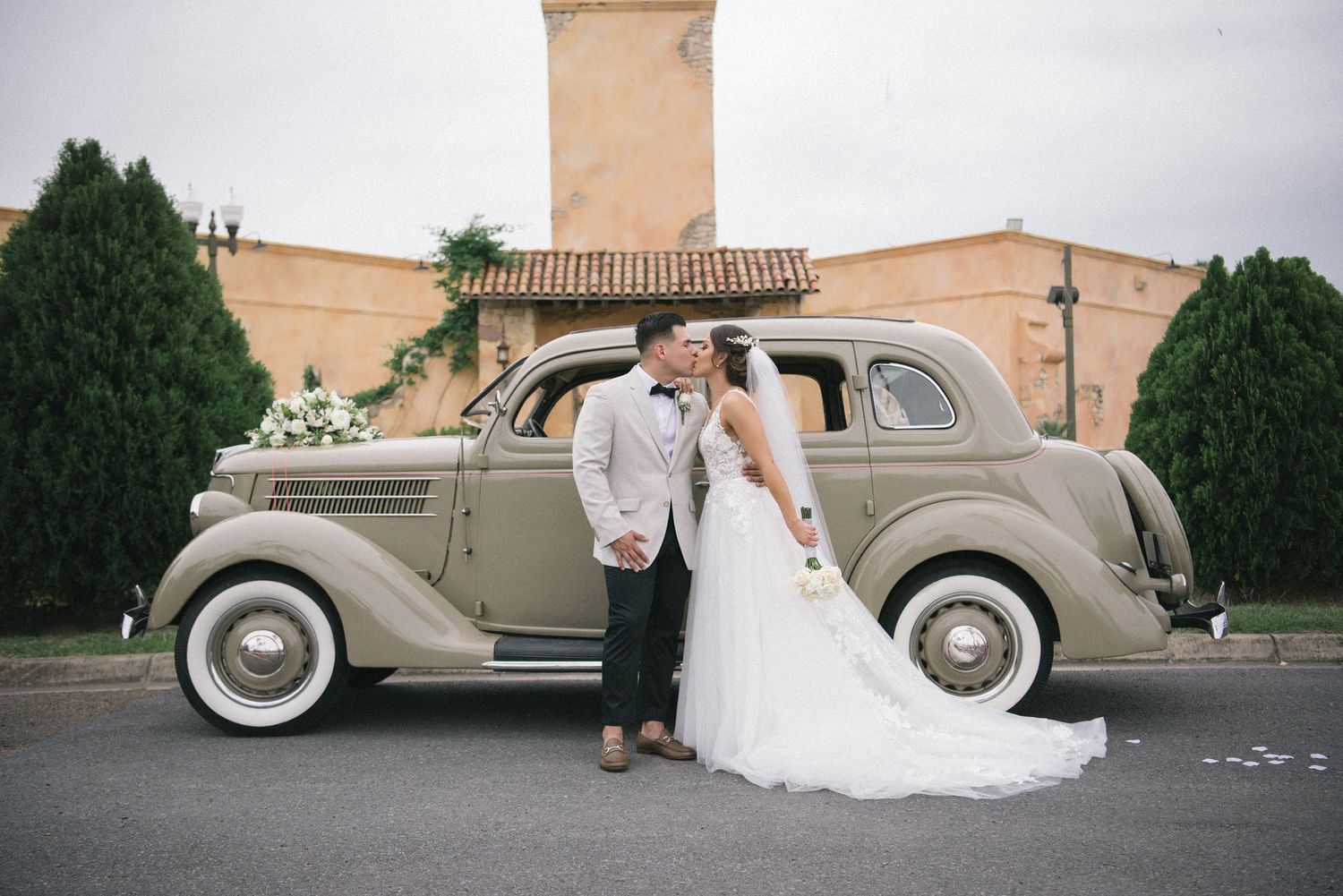 A couple poses with a classic vintage beige car at their wedding venue.