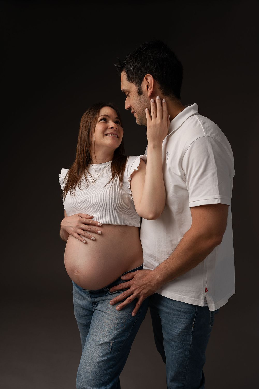 Intimate maternity portrait of couple in casual attire against dark background.