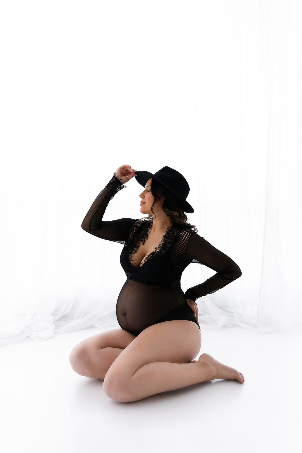 Stylish maternity pose in black bodysuit and hat against white backdrop.