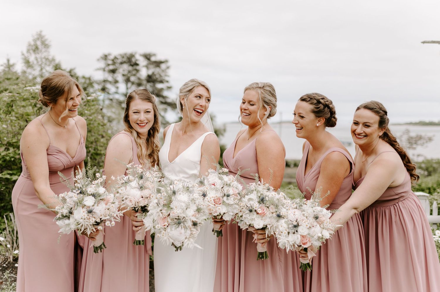 Bridesmaids in dusty pink dresses holding white floral bouquets stand together outdoors.