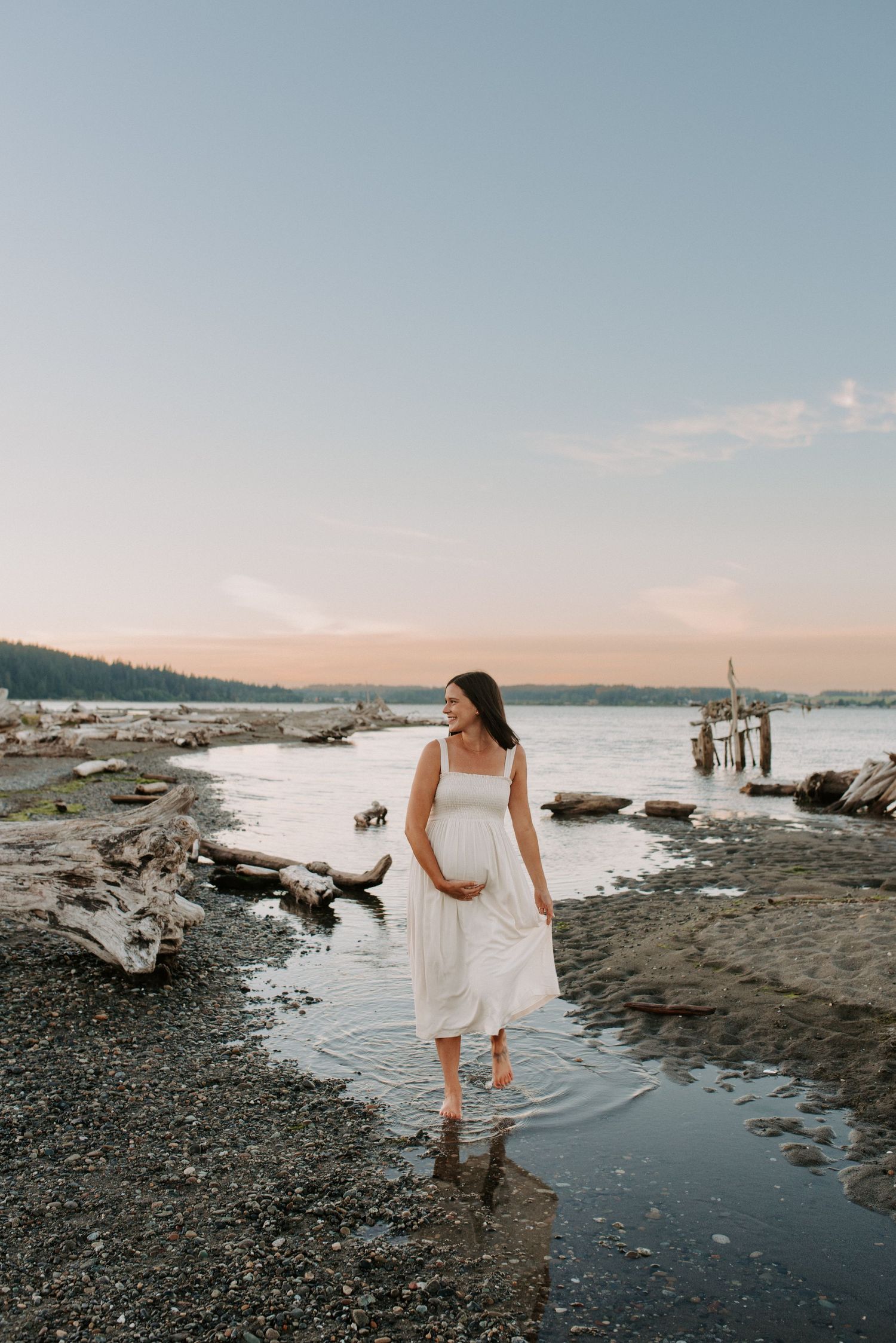Person in white dress walking along rocky beach shoreline at sunset with driftwood.