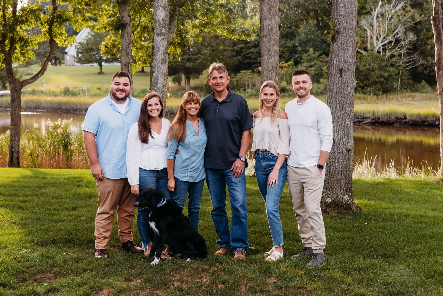 Group portrait taken outdoors by a pond with trees and natural landscape in background.