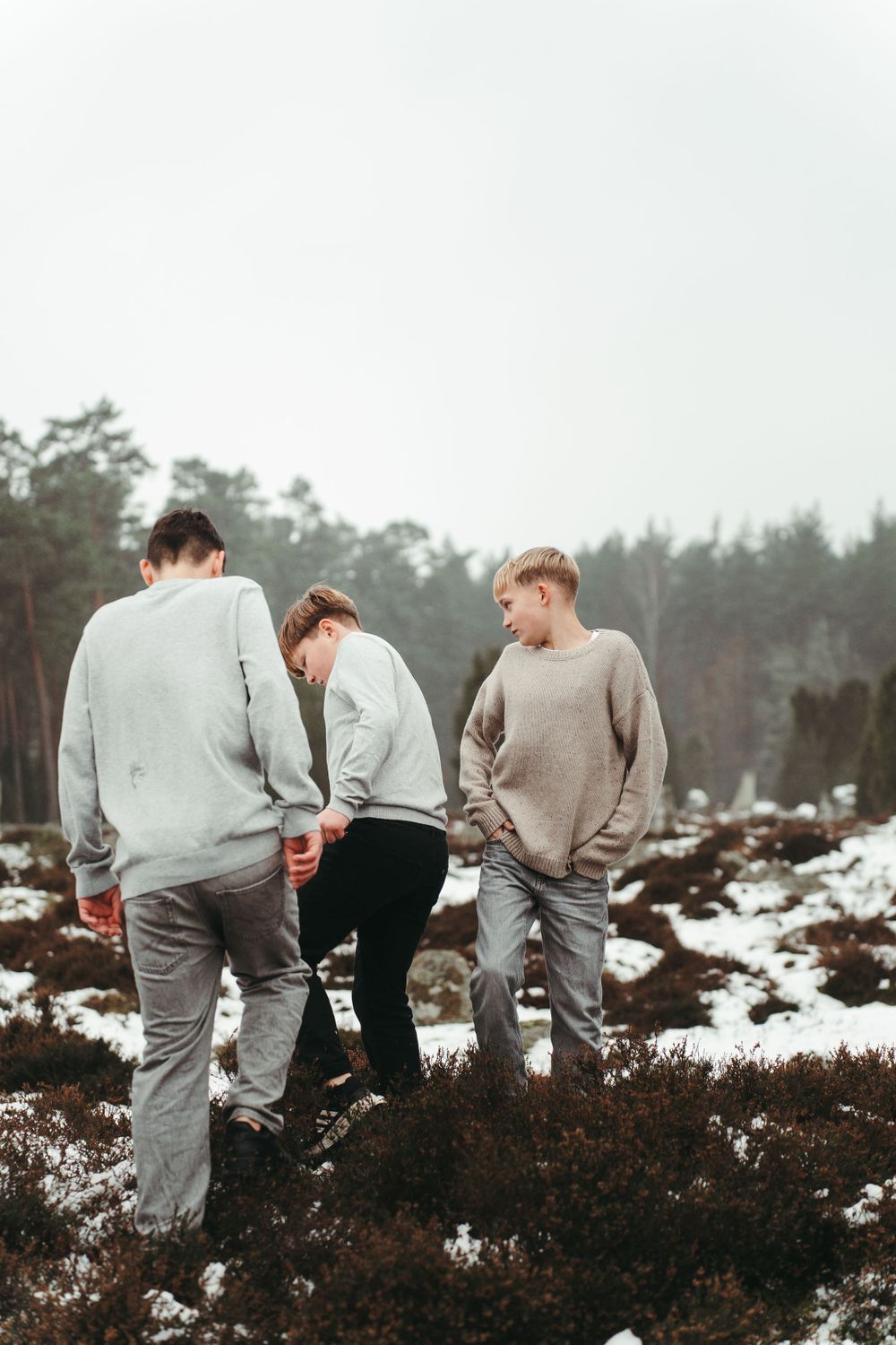 Three people in casual winter wear standing on snow-covered ground with pine trees in background.