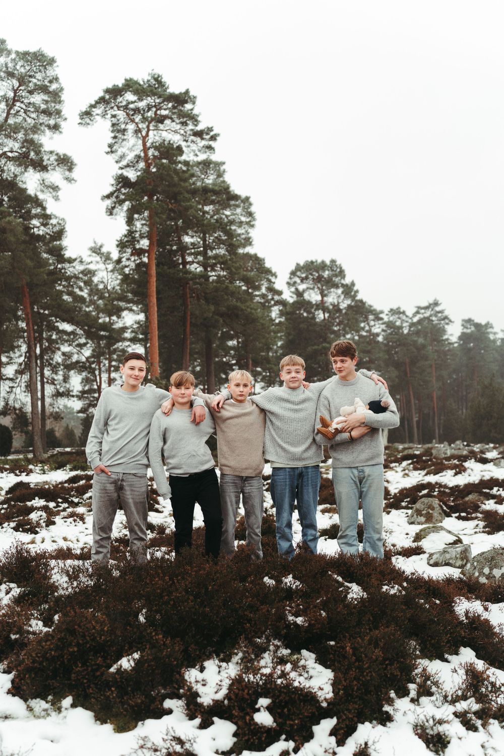 Group photo of friends in matching sweaters standing on snowy terrain with tall pine trees behind.