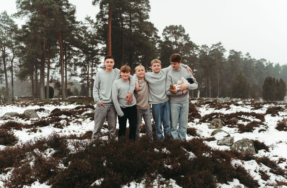 Friends in coordinated gray sweaters pose together on snowy ground with evergreen forest backdrop.