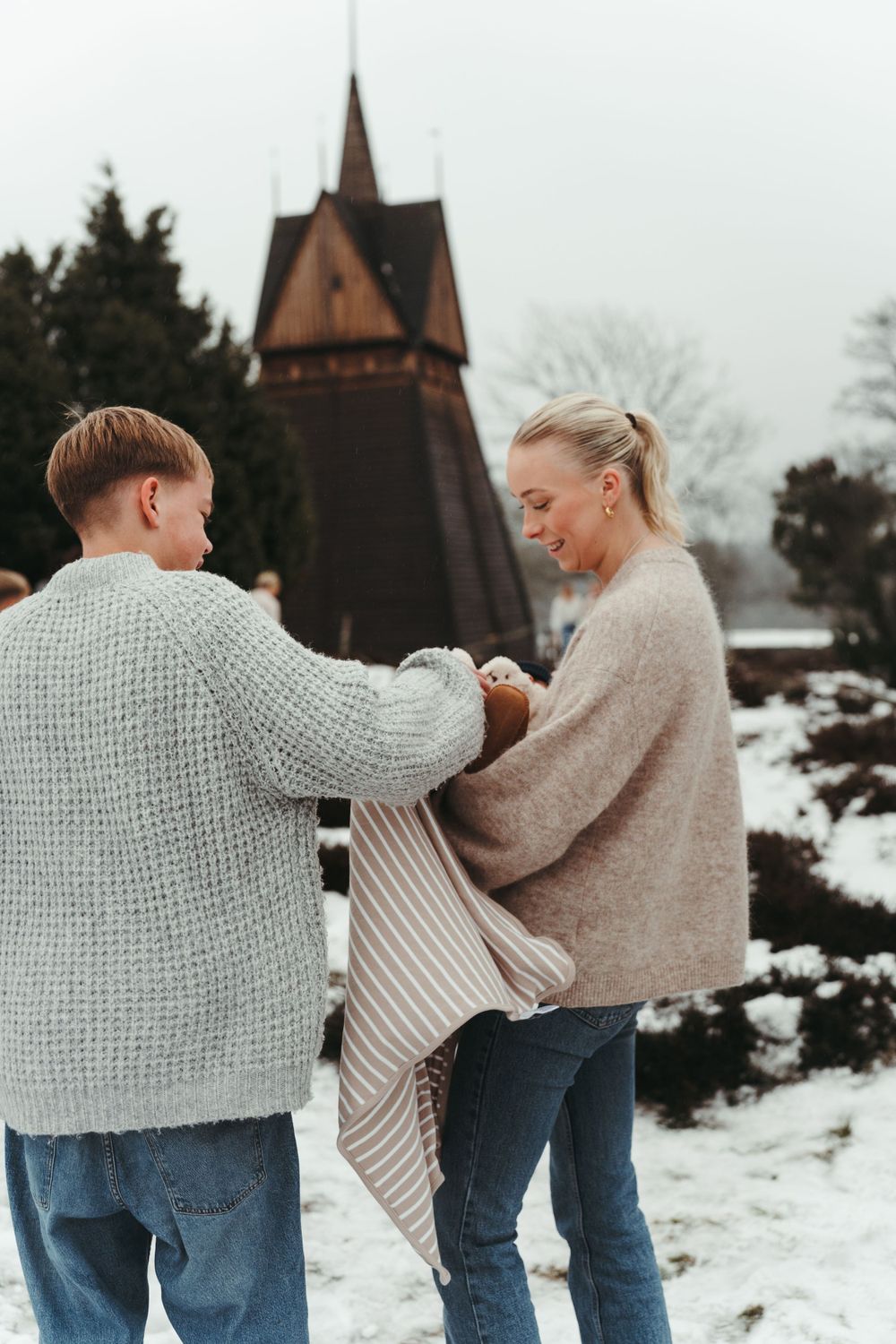 Two people hold a blanket together in front of a wooden church steeple on a snowy day.