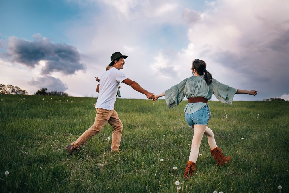 Two people run joyfully through a green meadow under dramatic cloudy skies.