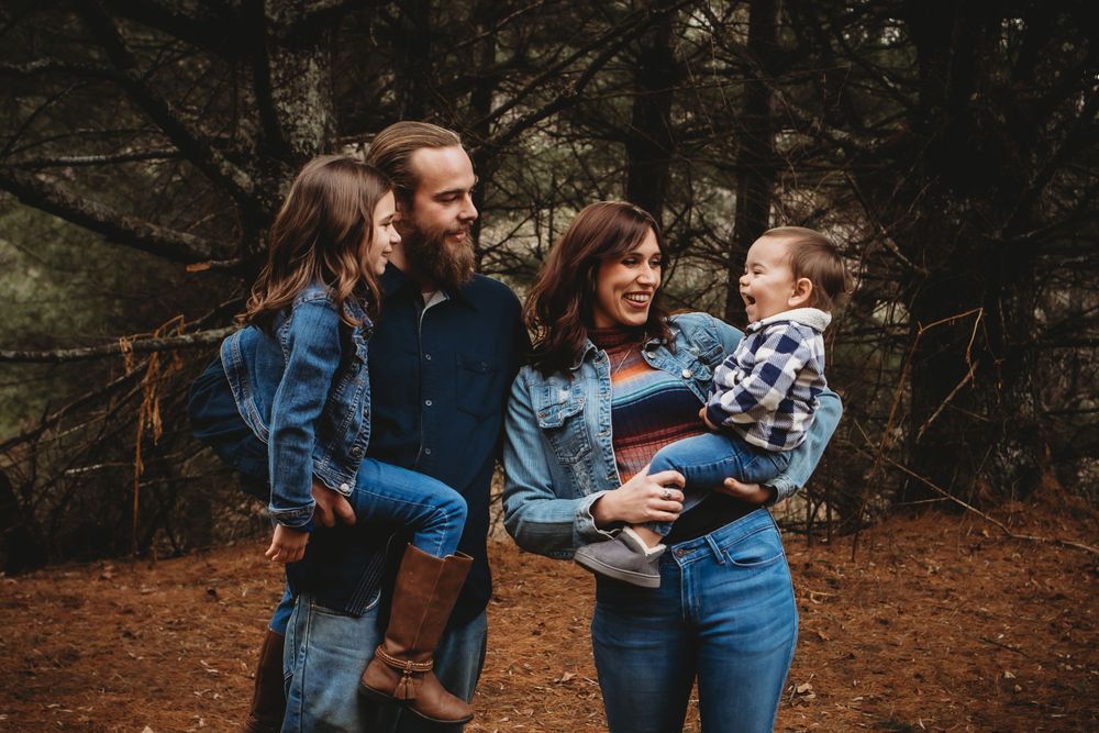 Family wearing coordinated denim outfits poses together in a wooded setting during autumn.
