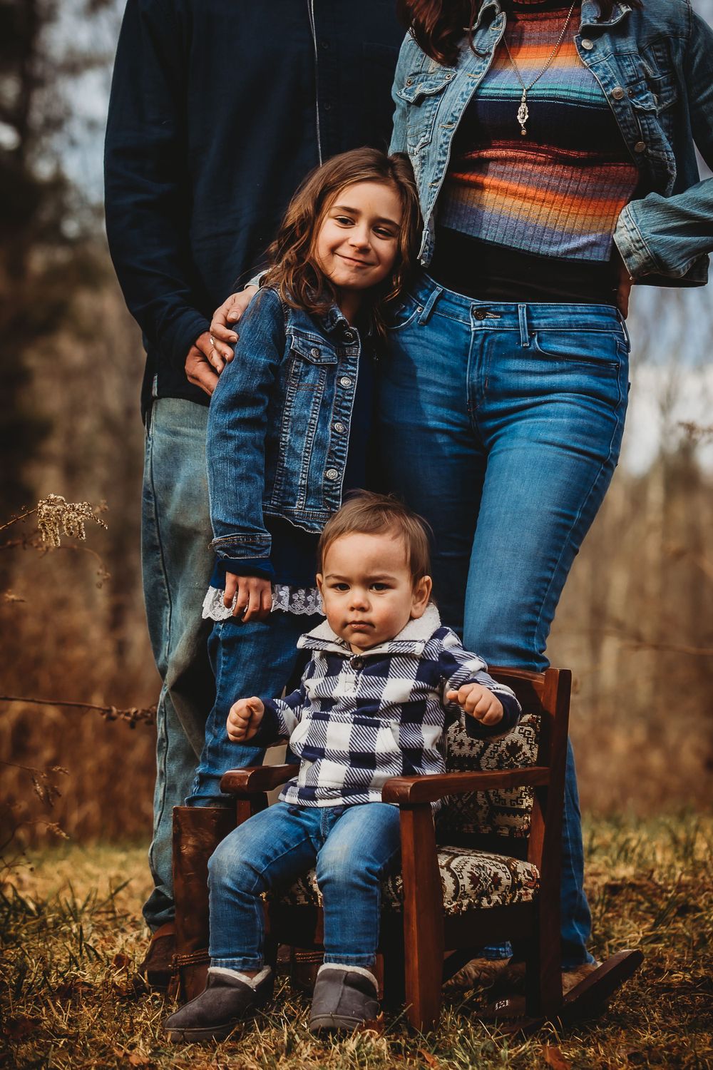 Casual family portrait with small child seated on vintage wooden chair outdoors.