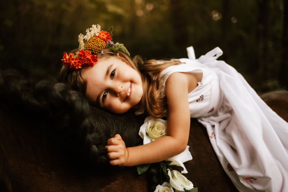 Child wearing flower crown and white dress smiles brightly in golden sunlight.
