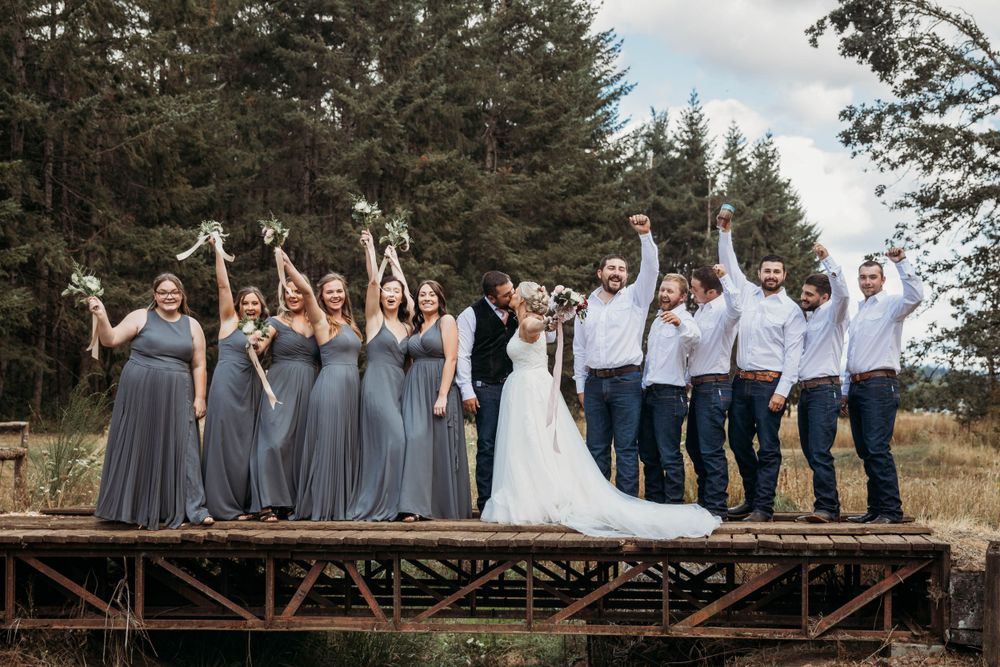 Wedding party poses playfully on a wooden bridge with bridesmaids in blue dresses.