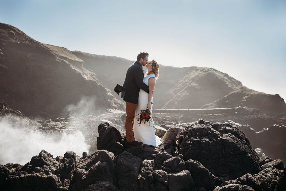 A romantic wedding couple poses along dramatic rocky cliffs by the ocean at sunset.