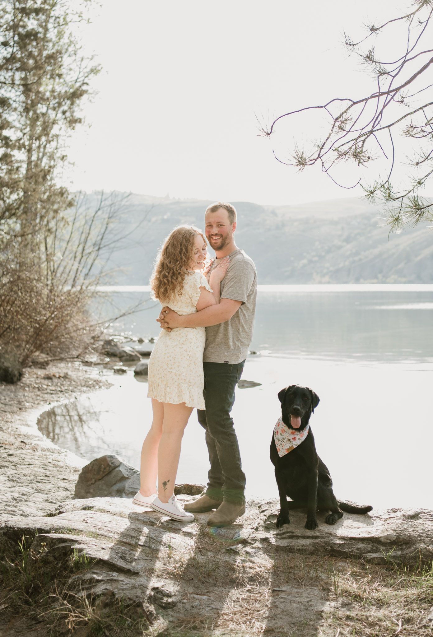 Kelowna engagement photo, couple embraces in front of Kal Lake in Vernon. They smile at their dog who is sitting beside