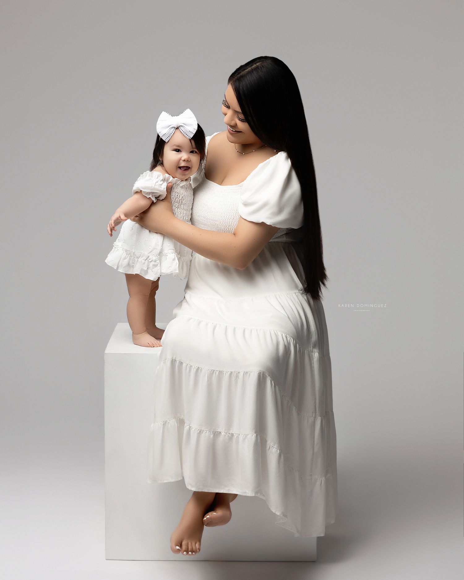 Mother and child wearing matching white dresses pose on white blocks in minimalist studio setting.