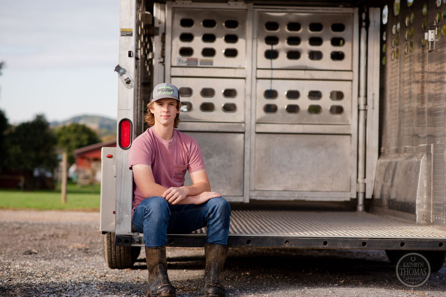 Person sits on back of livestock trailer wearing pink shirt and baseball cap.