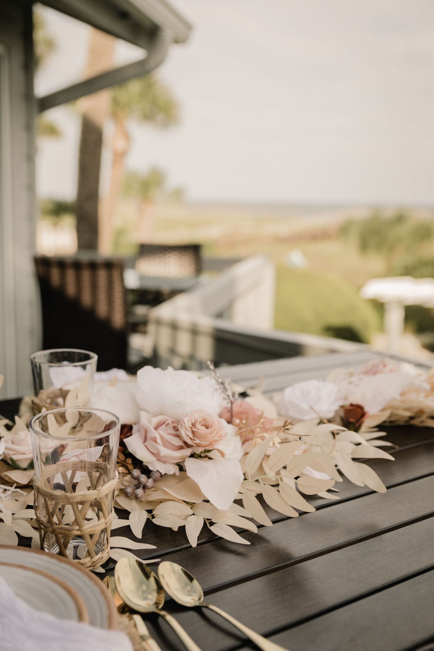 Outdoor wedding table setting with pink roses and palm leaves on dark wooden surface.