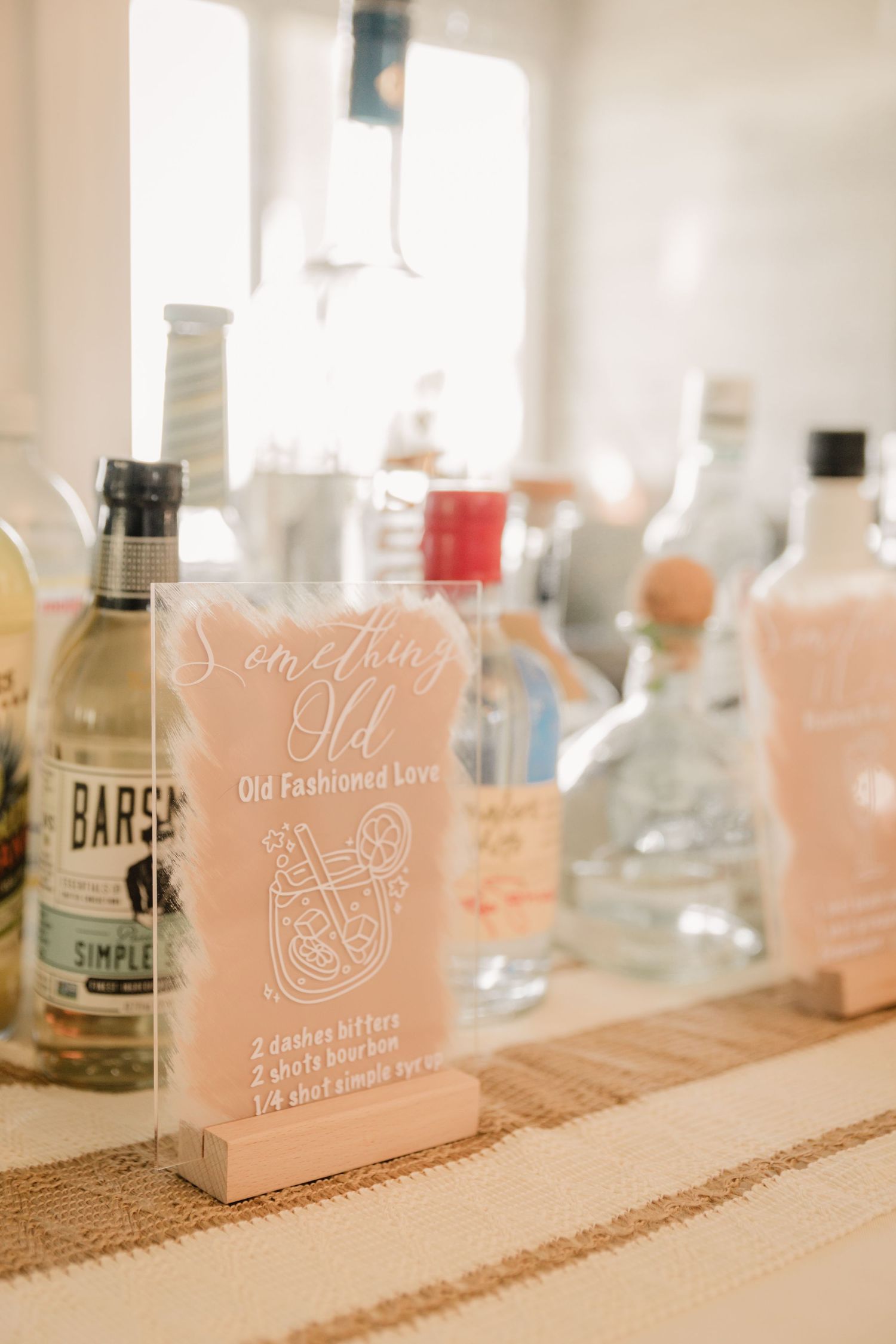 Close-up of wedding cocktail menu and assorted liquor bottles on burlap-lined shelf.