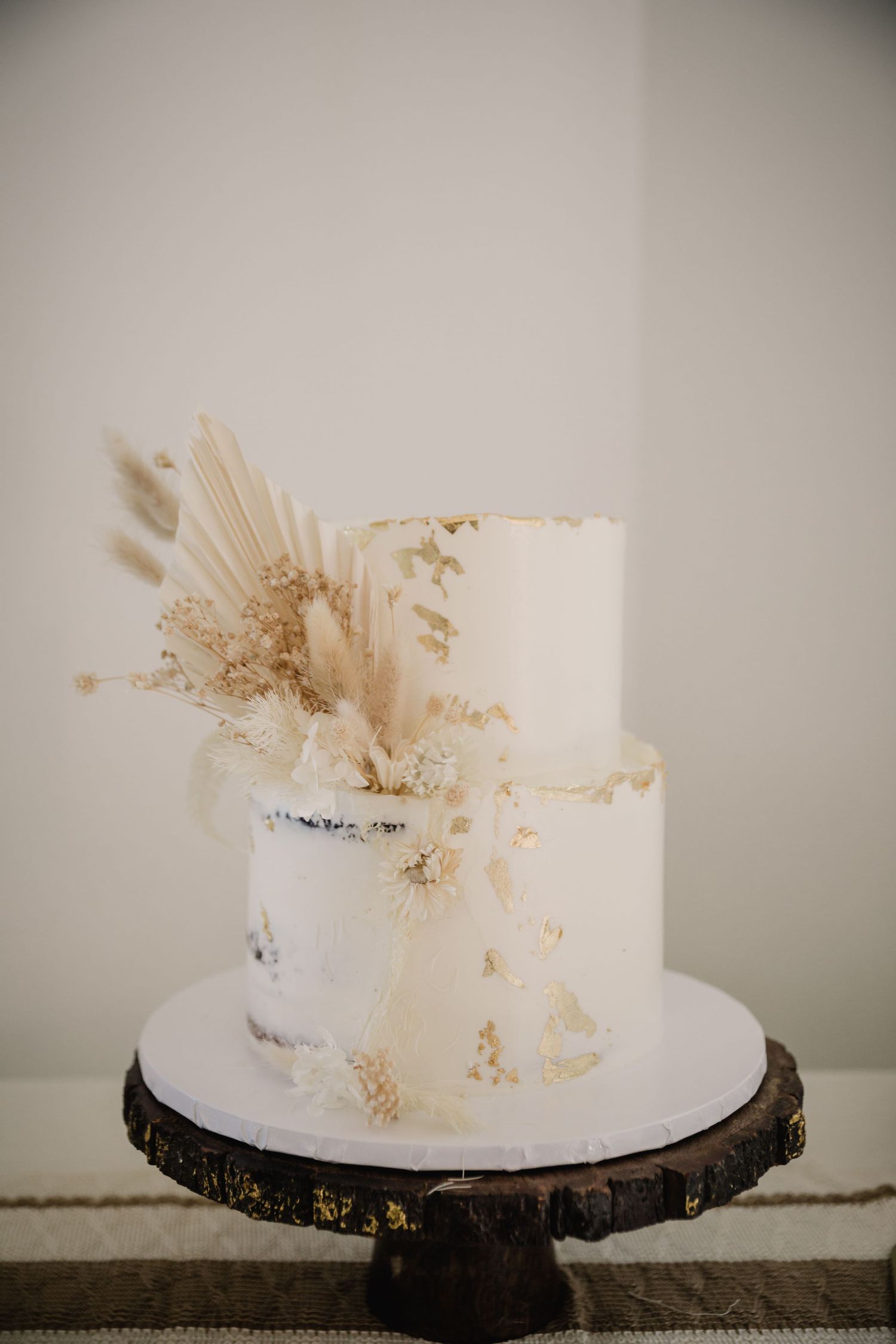 Elegant two-tier white wedding cake with dried pampas grass and gold leaf accents displayed on wooden cake stand.