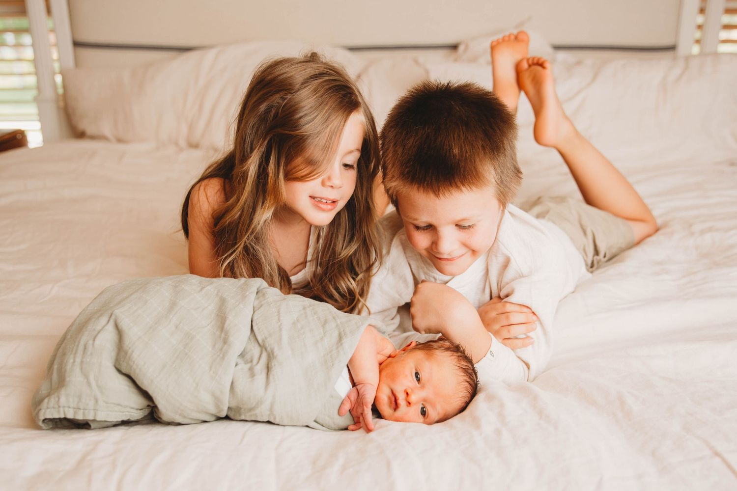 Children lying on white bed looking at sleeping newborn brother in soft lighting.