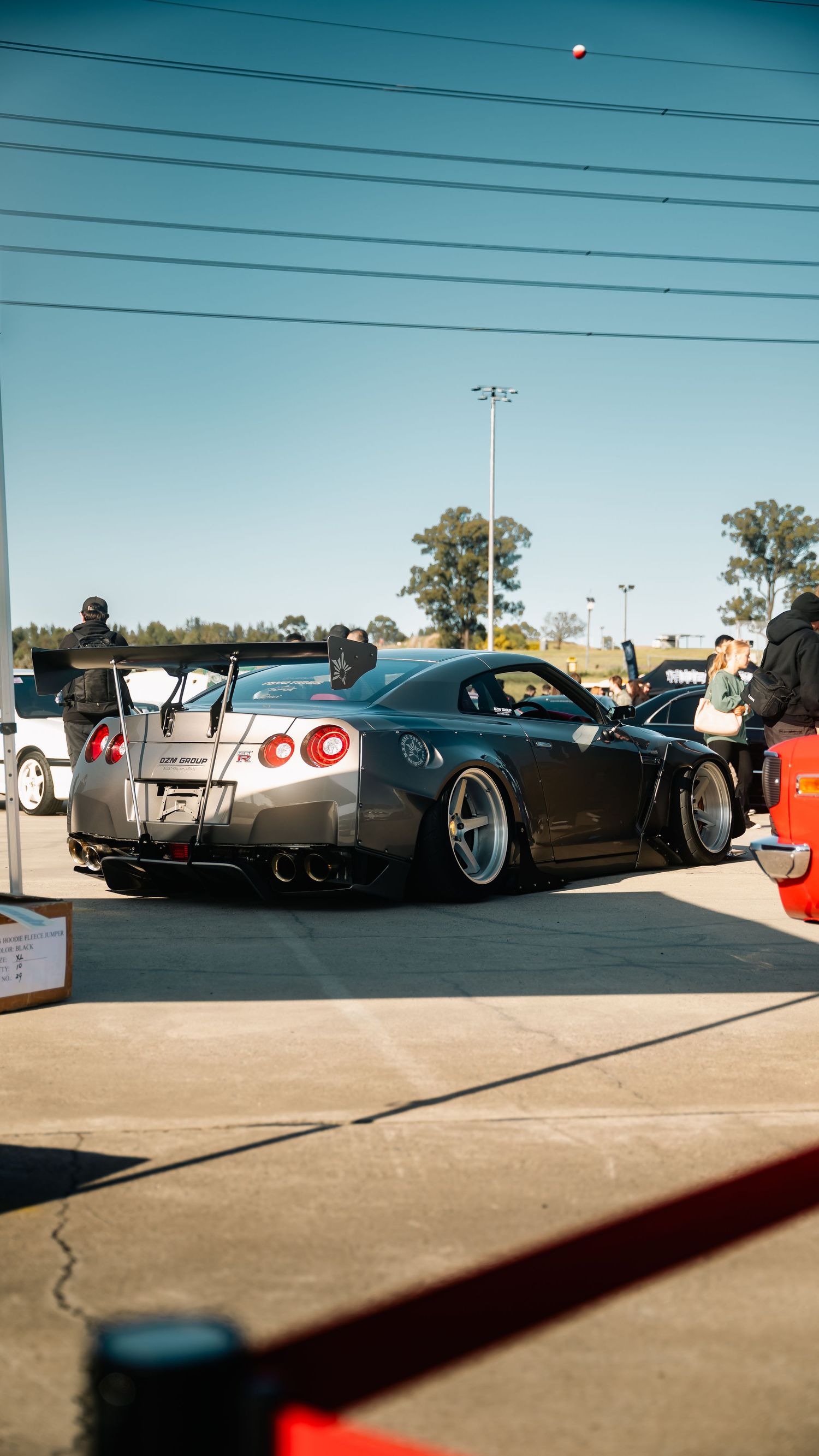 Modified Nissan GTR with custom wheels and body kit parked at a car show under power lines.