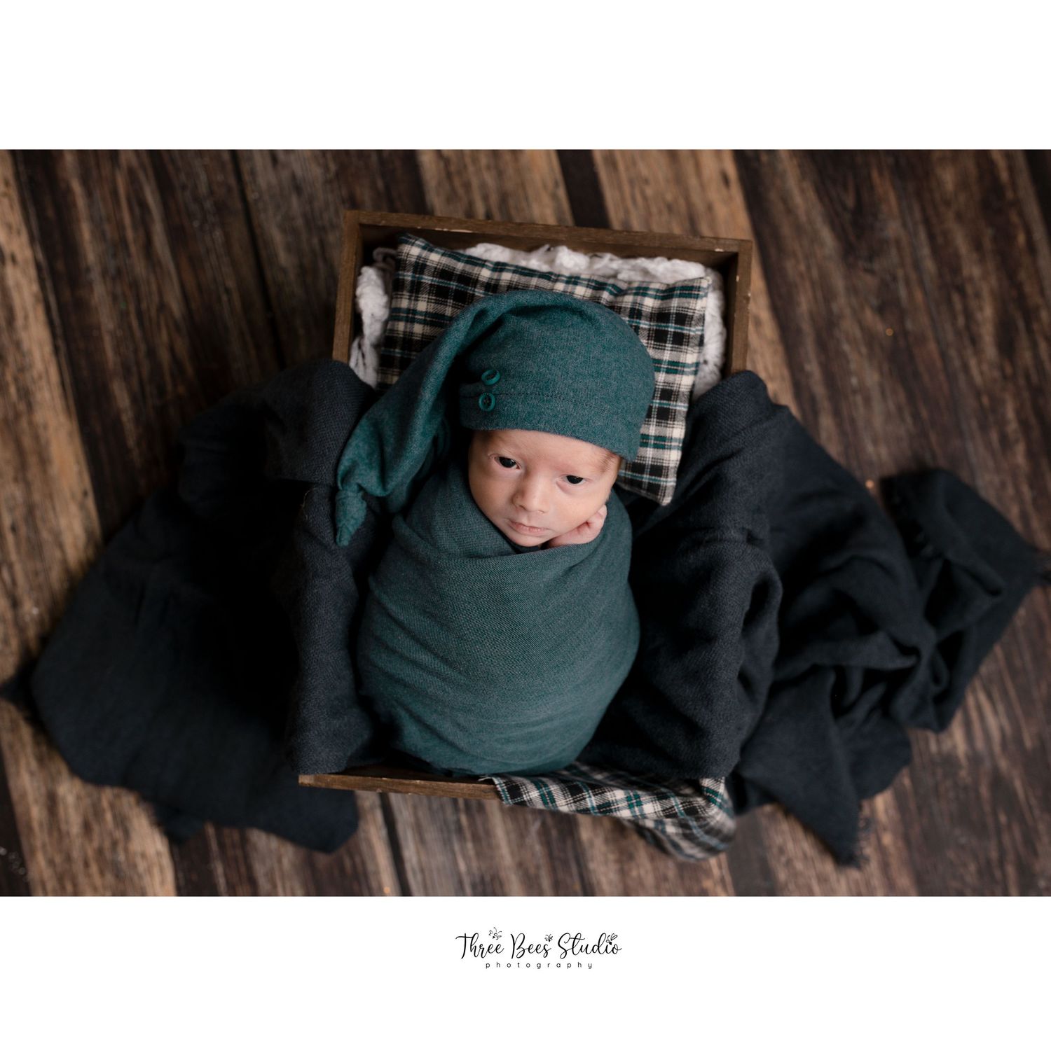 Newborn wrapped in teal swaddle on plaid pillow against dark wooden background.
