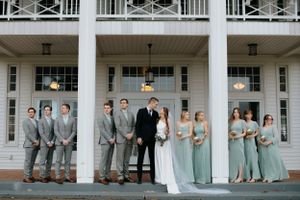 Wedding party in gray suits and sage green dresses posed on white columned porch.