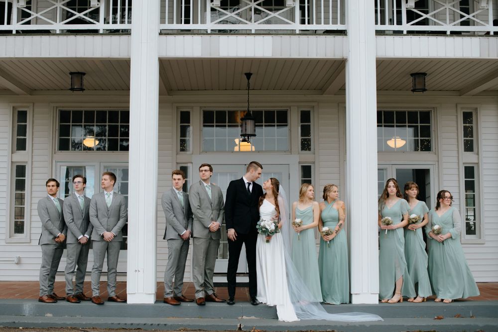Wedding party in gray suits and sage green dresses posed on white columned porch.