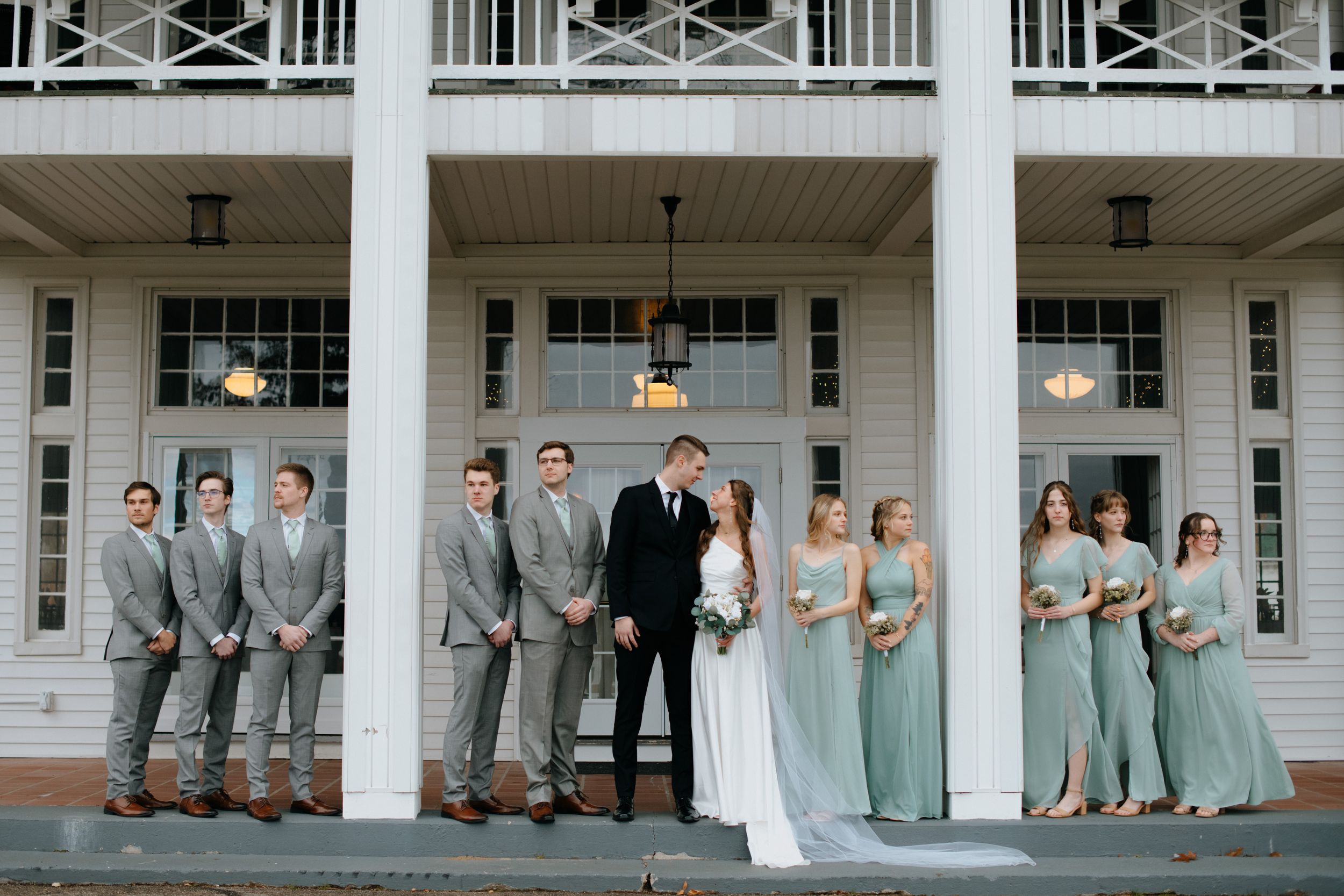 Wedding party in gray suits and sage green dresses posed on white columned porch.