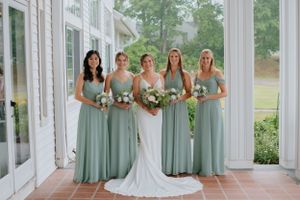 Bridal party in sage green bridesmaid dresses poses in sunlit conservatory.