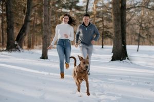 Couple running in snowy forest with dog on winter day.