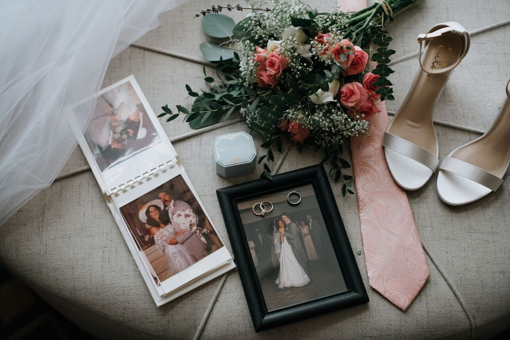 Wedding details flatlay with bouquet, photographs, shoes and pink tie on beige surface.