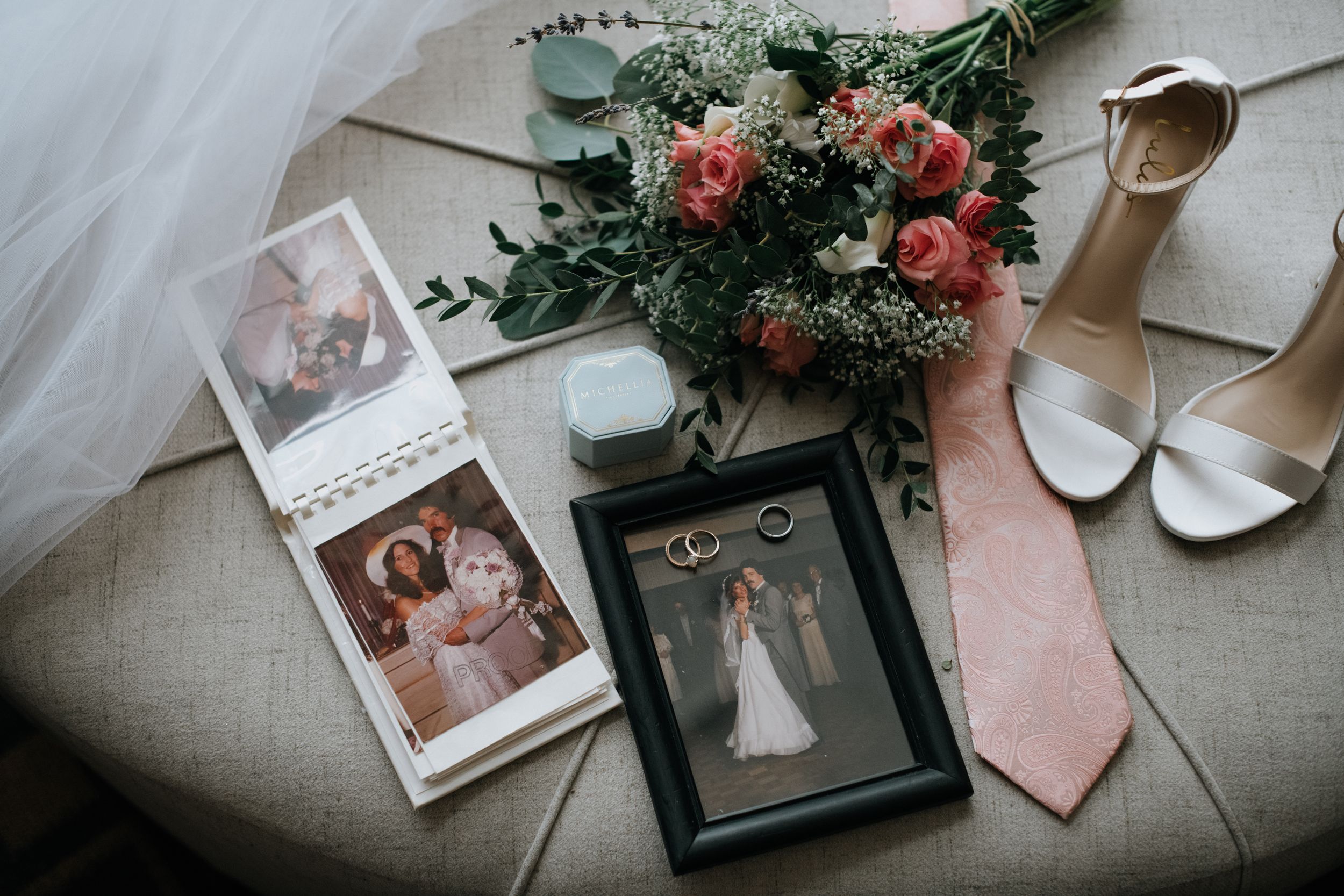 Wedding details flatlay with bouquet, photographs, shoes and pink tie on beige surface.