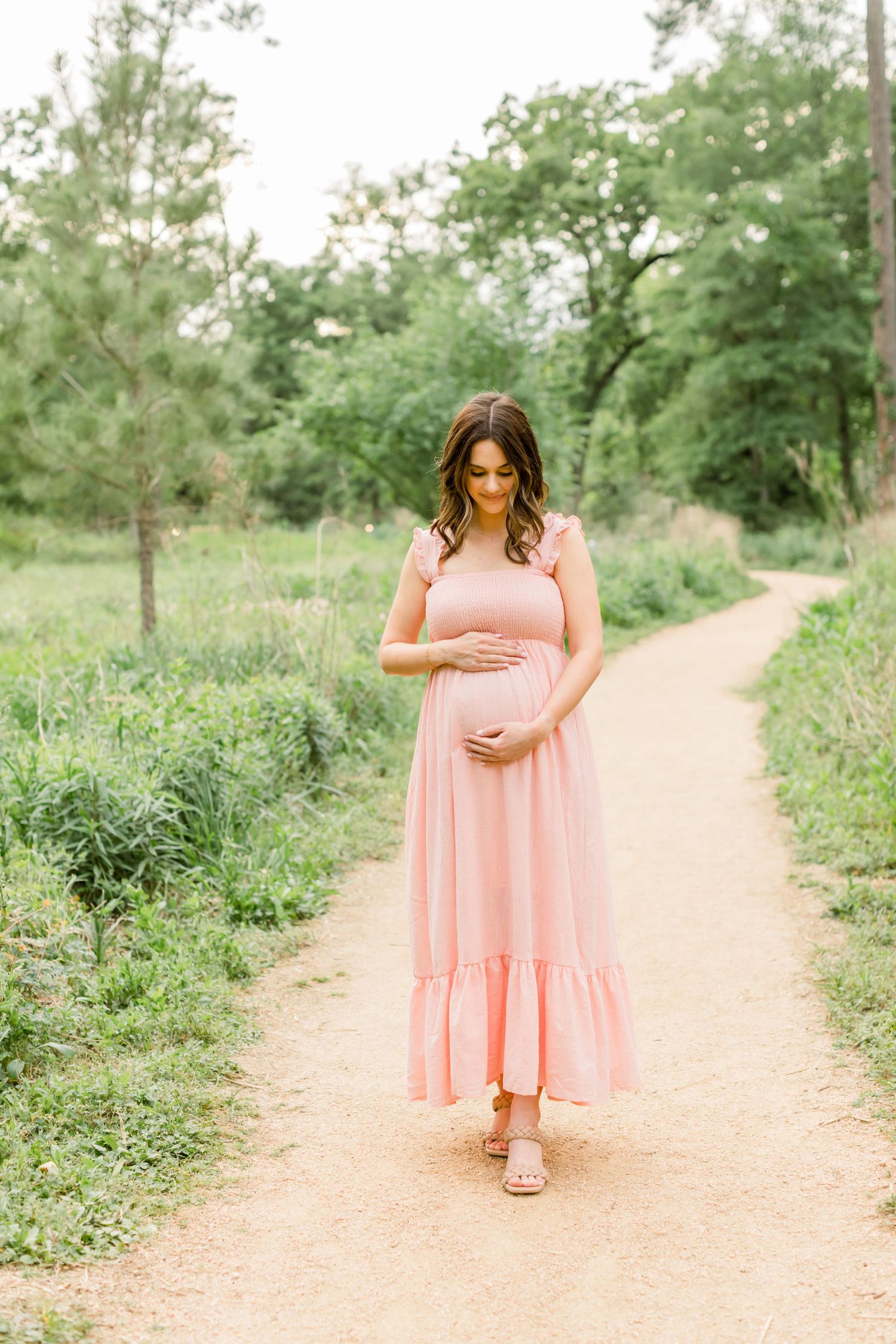 A woman in a blush pink tiered maxi dress stands on a winding dirt trail in a park.