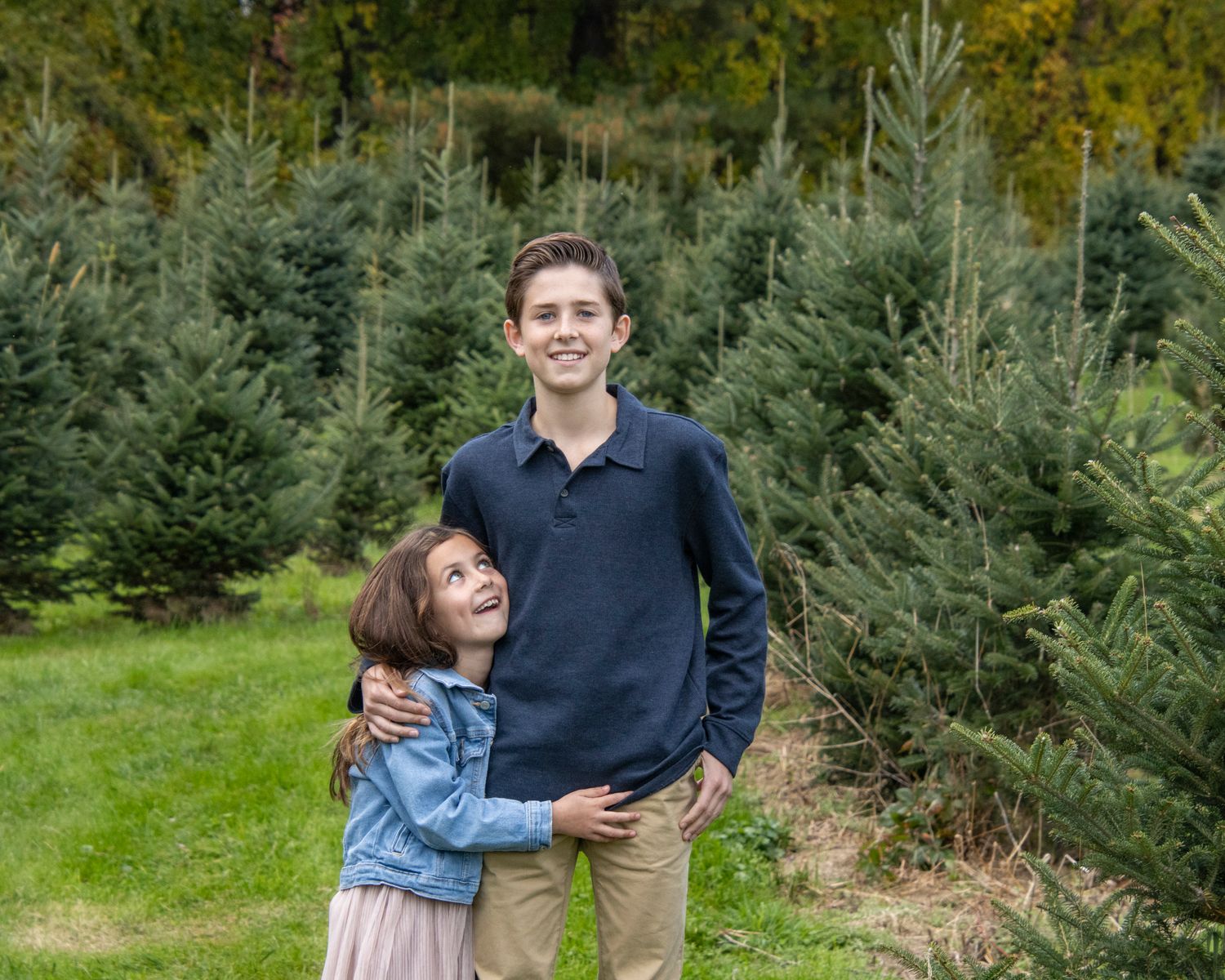Two siblings share a joyful moment together among evergreen trees at a Christmas tree farm.