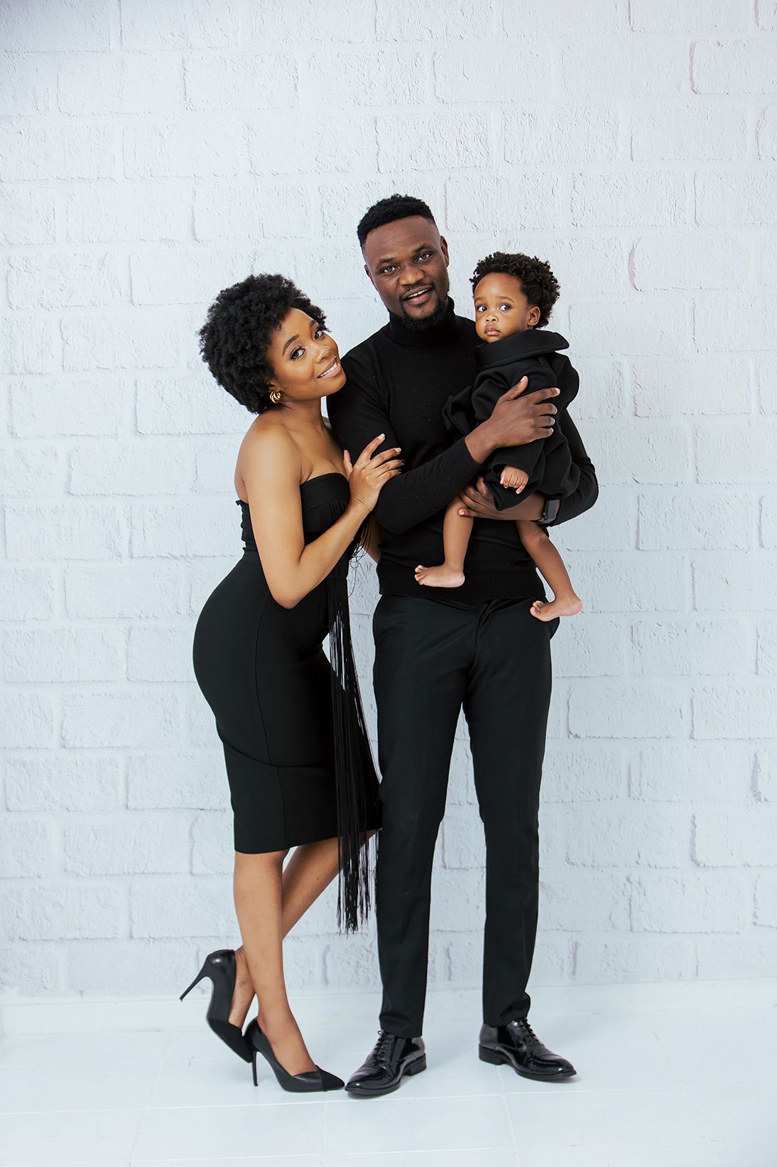 A stylish family portrait in matching black outfits against a white brick wall background.