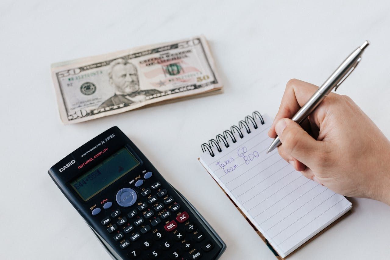 Calculator, ten dollar bill, and hand writing in notepad on white surface.