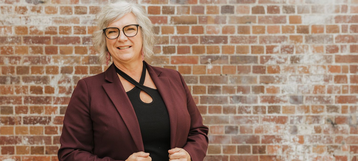 Professional wearing burgundy blazer and glasses poses against rustic brick wall background.
