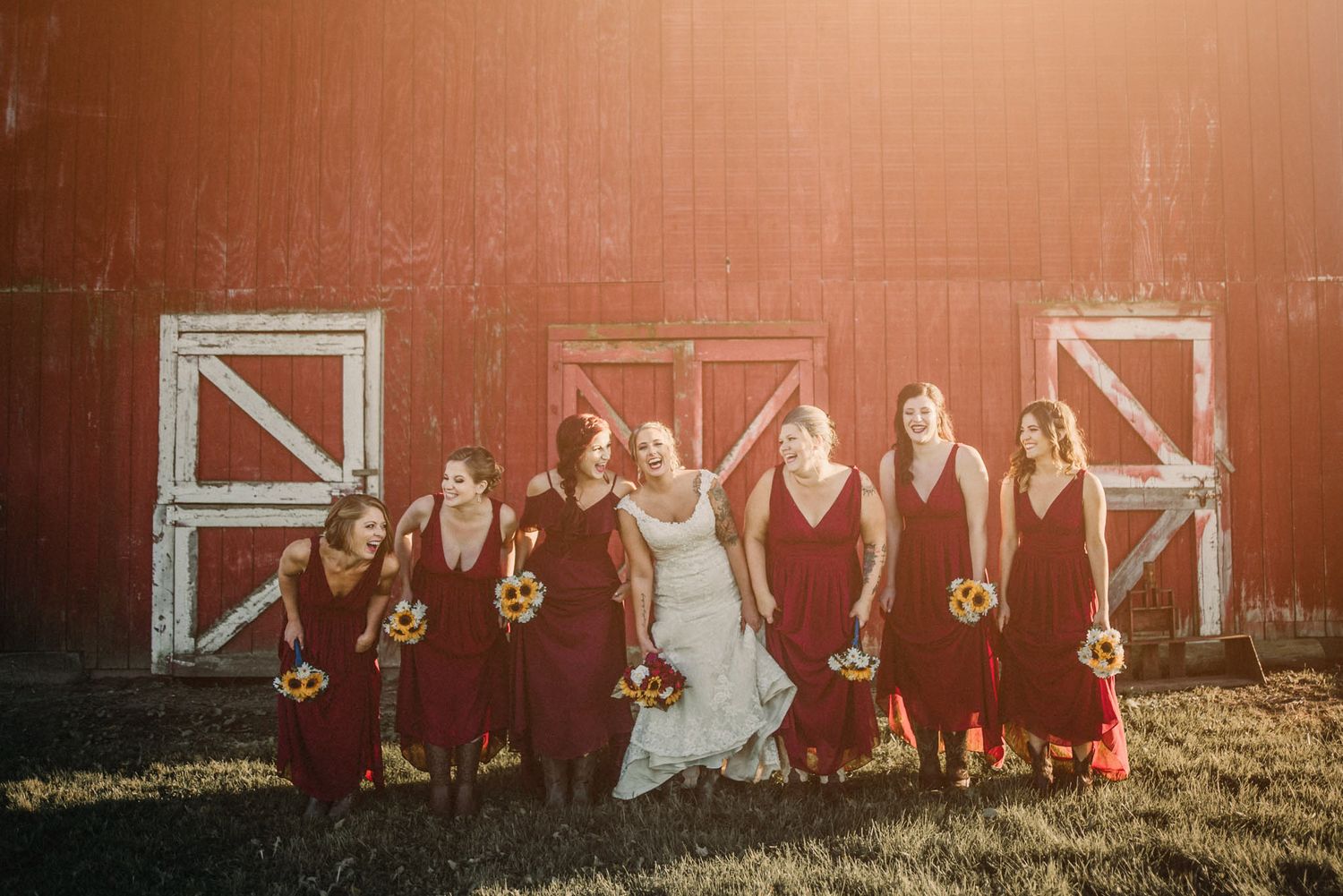 Wedding party in burgundy dresses and white gown pose playfully against a rustic red barn at sunset.