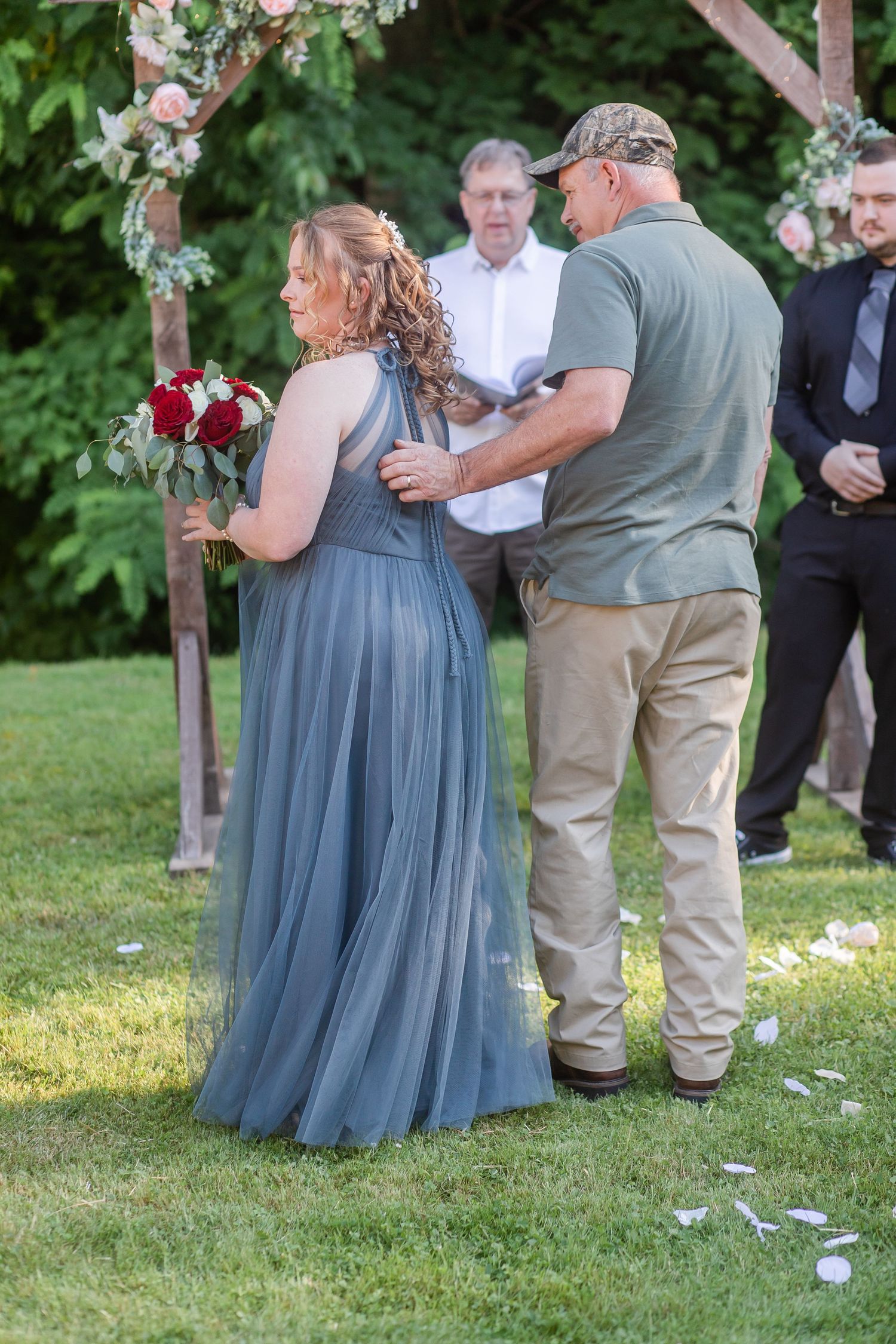 Outdoor wedding ceremony with bridesmaids in dusty blue dresses and floral arch decorations.