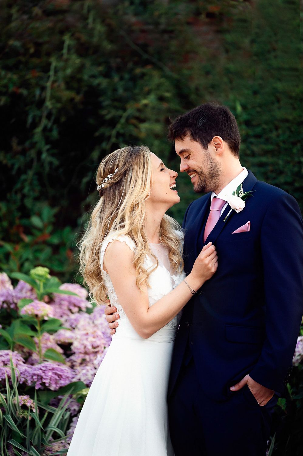 A romantic wedding photo of a couple embracing outdoors with pink flowers in the background.