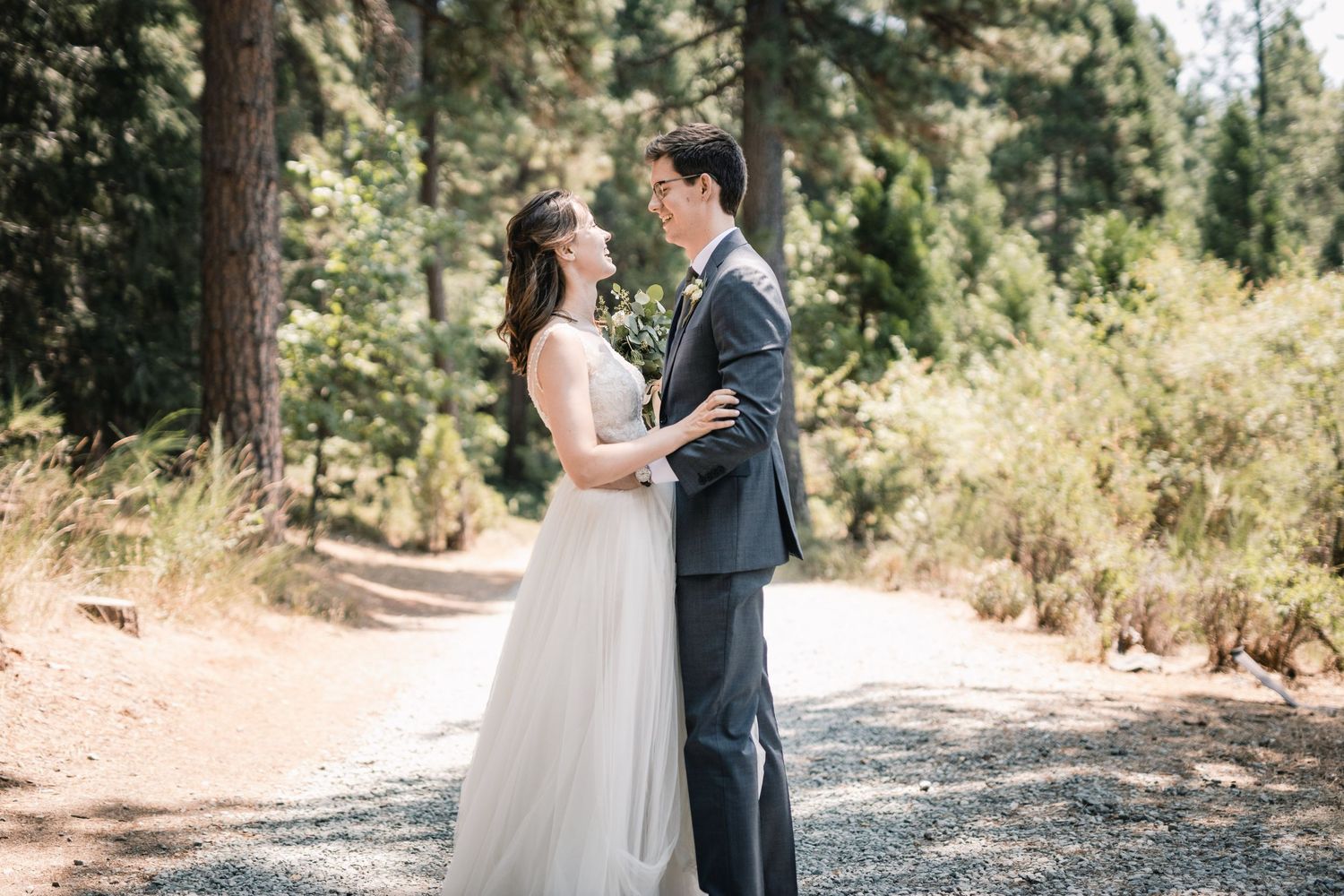 Newlyweds embrace on a forest path wearing wedding attire during their outdoor ceremony.