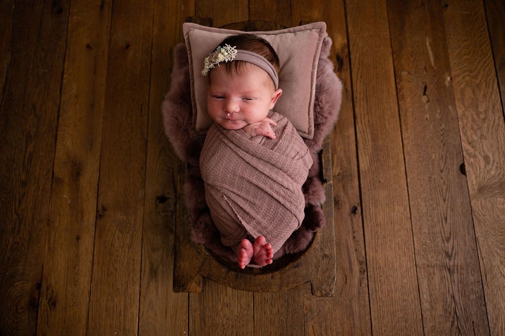 A sleeping newborn wrapped in mauve fabric rests in wooden prop bowl on hardwood floor.