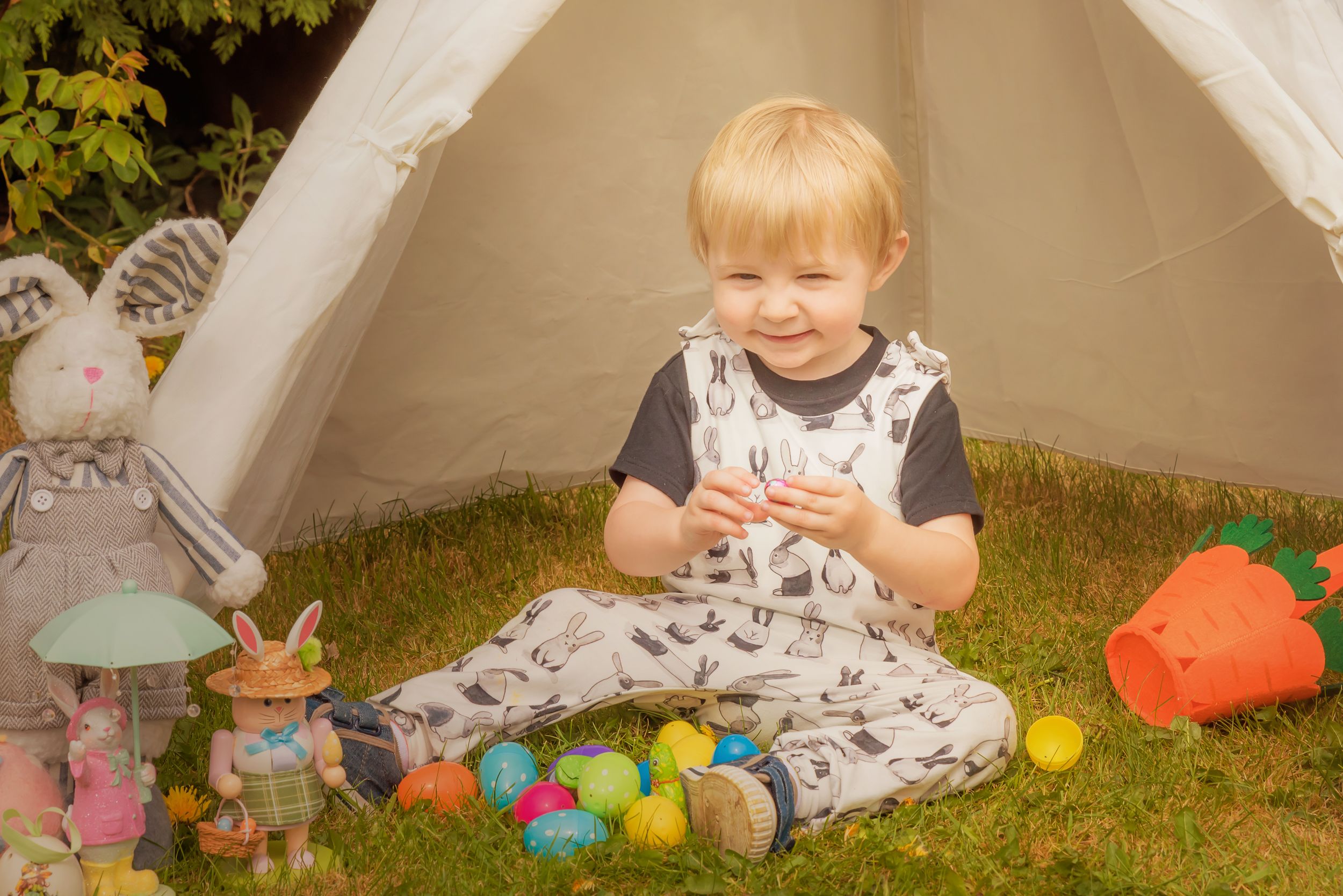 A child sits on grass surrounded by colorful toys under a white tent.