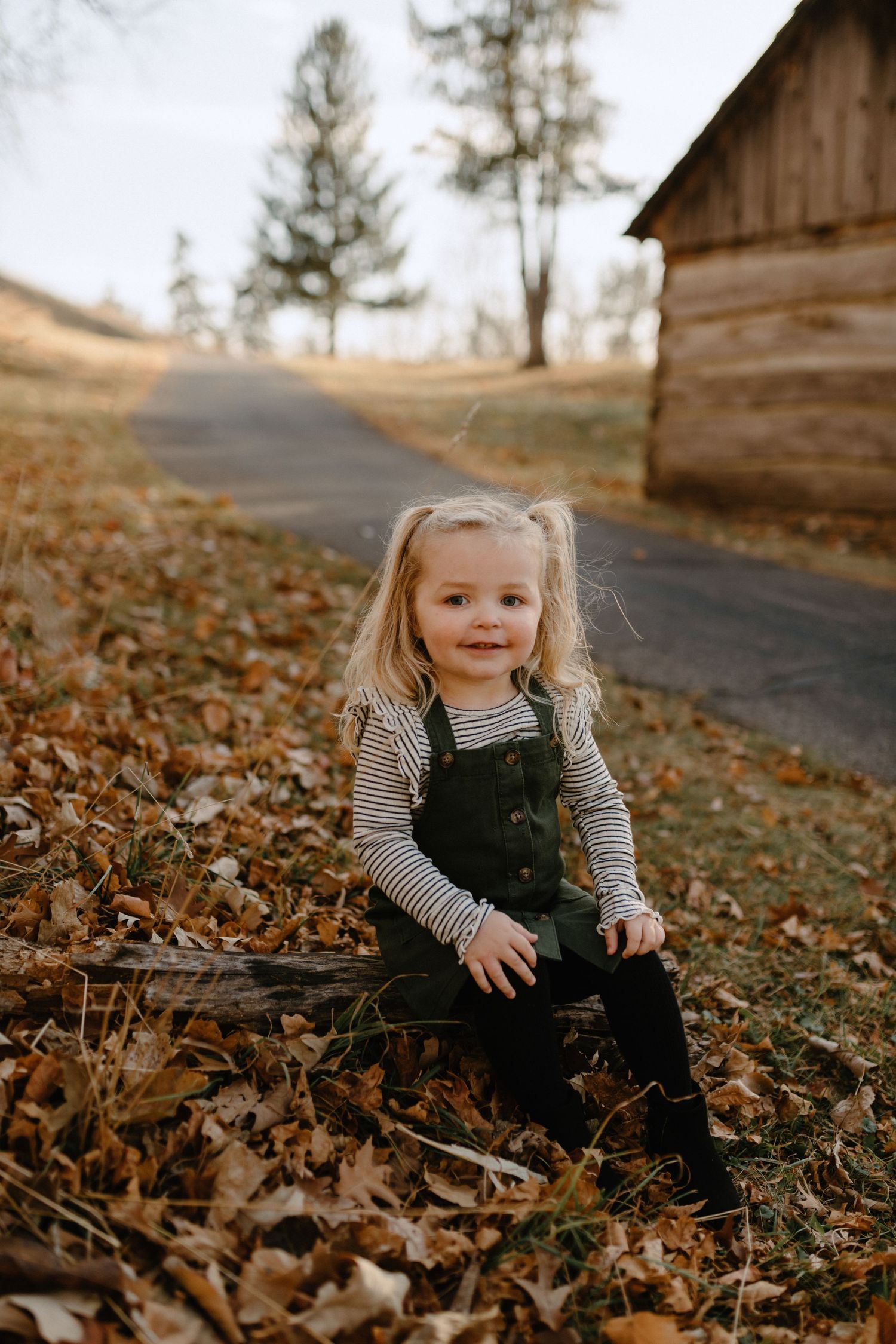 Child in striped shirt and overalls sitting among fall leaves near rustic wooden building.