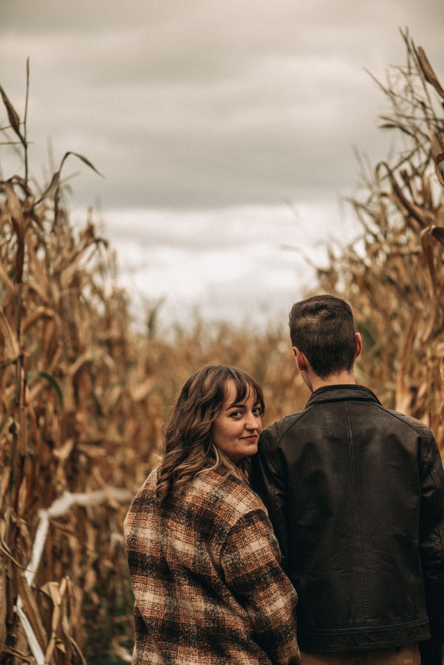 A couple walks through a golden cornfield on a moody autumn day wearing plaid and dark jackets.