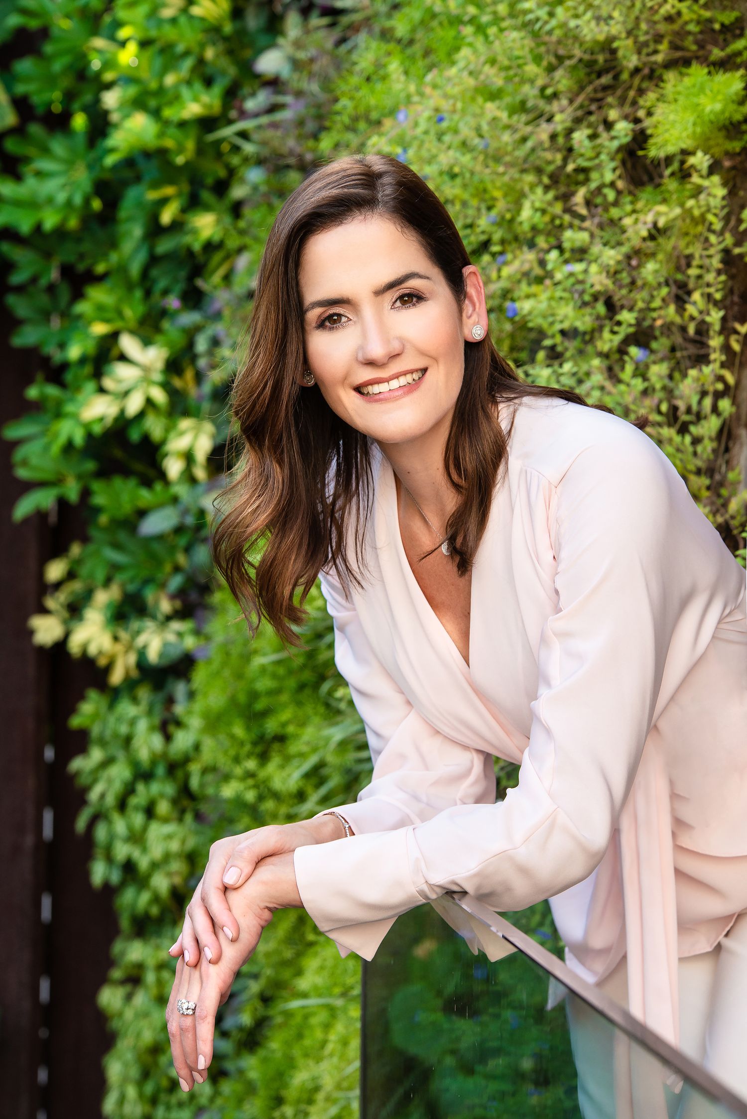 A casual portrait against green foliage and brick wall wearing a light pink wrap top.