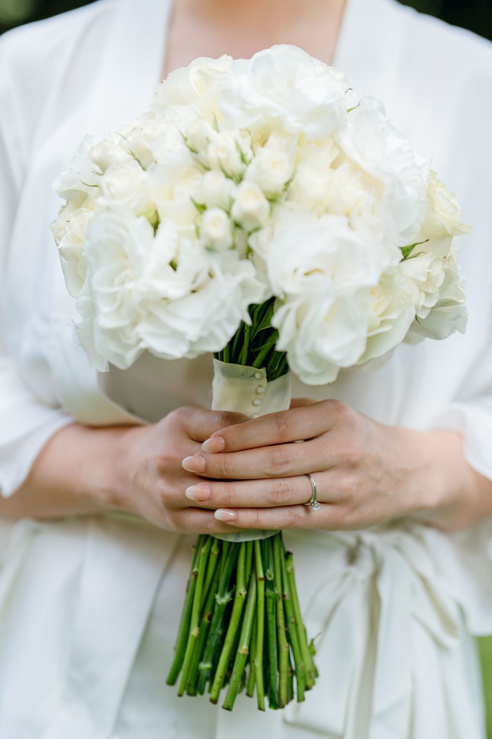 A classic white rose wedding bouquet with long green stems held against a white dress.