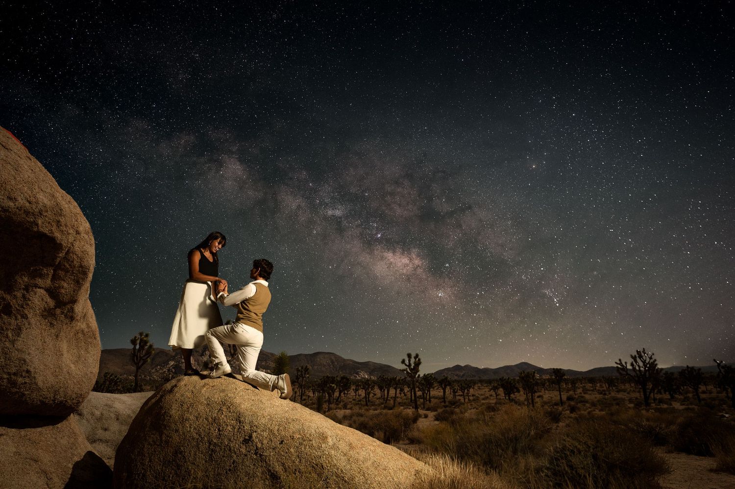 Nighttime engagement photo with couple on boulder under starlit sky in Joshua Tree.