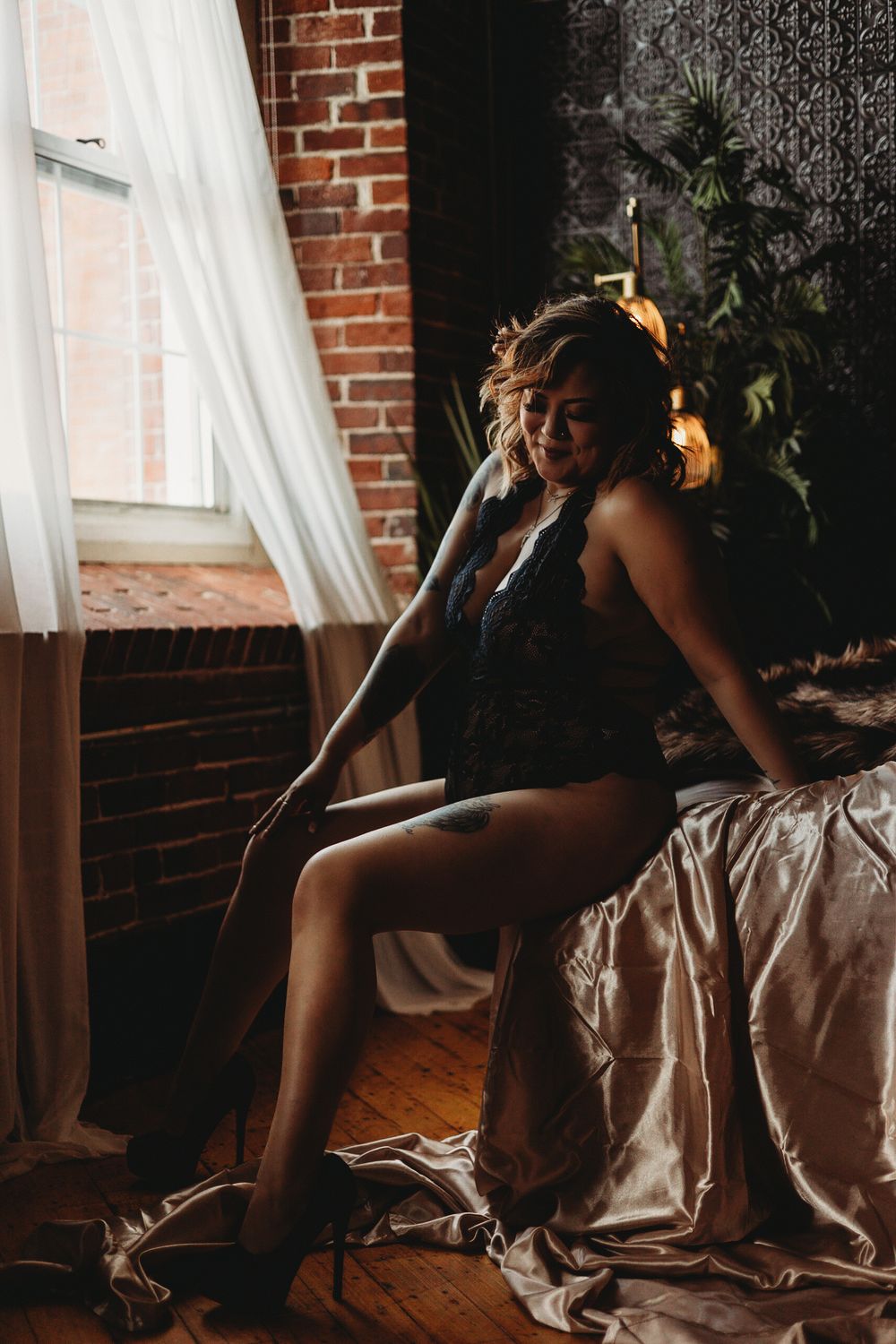 Dramatic boudoir portrait by brick wall with natural window light and flowing curtains.