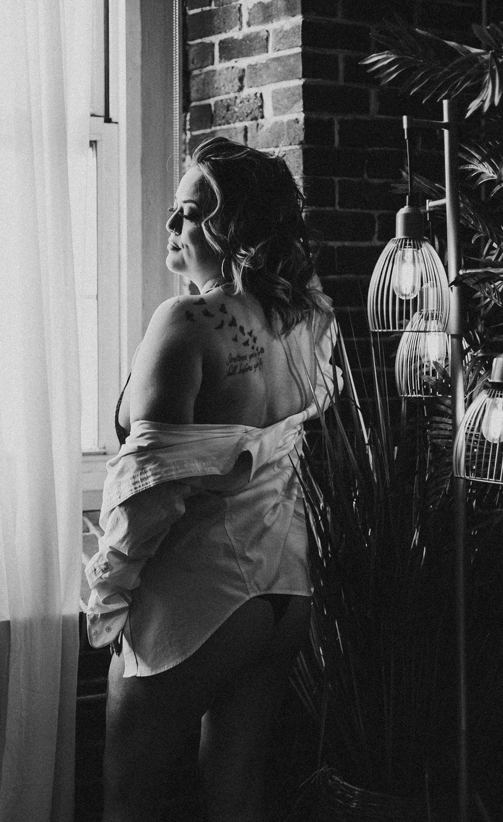 Monochrome portrait by window with exposed shoulder and hanging lanterns in background.
